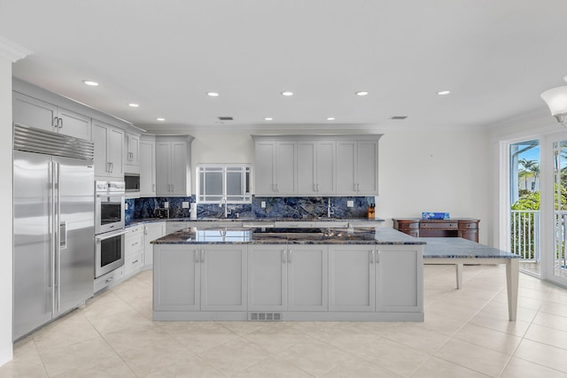 kitchen with gray cabinetry, dark stone countertops, built in appliances, a kitchen island, and decorative backsplash