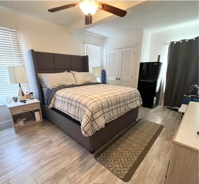 bedroom featuring ceiling fan, light wood-style floors, a closet, freestanding refrigerator, and beamed ceiling