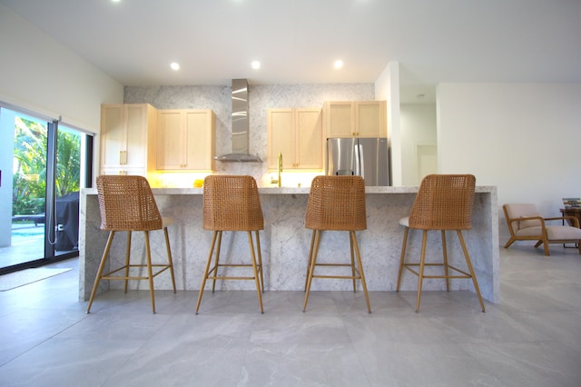 kitchen with a kitchen bar, stainless steel fridge, wall chimney exhaust hood, and light brown cabinets