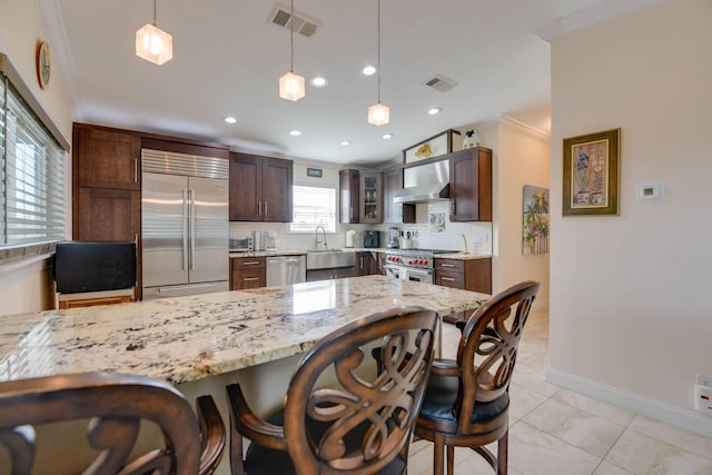 kitchen with high end appliances, decorative light fixtures, wall chimney range hood, light stone countertops, and backsplash