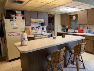 kitchen featuring light tile patterned floors, white refrigerator, range with electric stovetop, a kitchen breakfast bar, and a drop ceiling