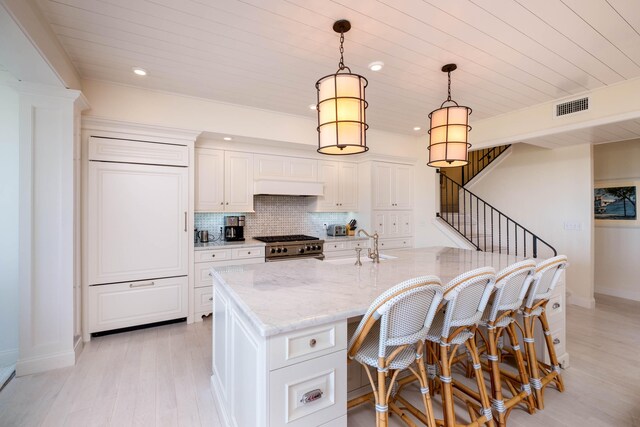 kitchen with pendant lighting, high end stainless steel range, light stone countertops, white cabinets, and a center island with sink