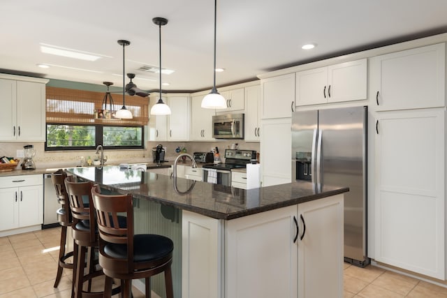 kitchen featuring light tile patterned flooring, appliances with stainless steel finishes, dark stone counters, hanging light fixtures, and a center island with sink