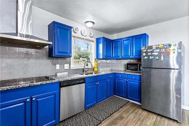 kitchen featuring light stone counters, appliances with stainless steel finishes, a sink, wall chimney range hood, and blue cabinets