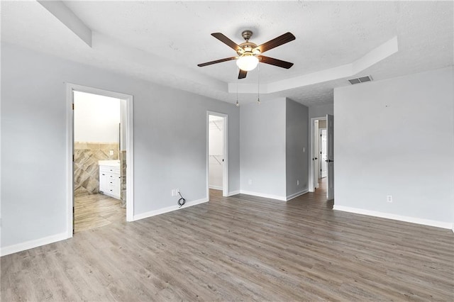 unfurnished bedroom with a walk in closet, ensuite bath, ceiling fan, a raised ceiling, and hardwood / wood-style floors