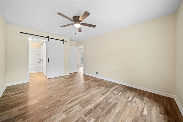 unfurnished bedroom with light wood-type flooring, a barn door, ensuite bath, and ceiling fan