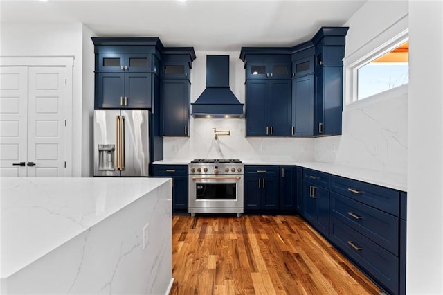 kitchen with wood finished floors, blue cabinets, premium appliances, wall chimney range hood, and decorative backsplash