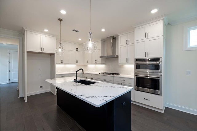 kitchen with wall chimney exhaust hood, a kitchen island with sink, sink, white cabinets, and hanging light fixtures