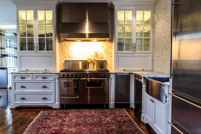 kitchen featuring light stone counters, stainless steel appliances, wall chimney exhaust hood, tasteful backsplash, and glass insert cabinets