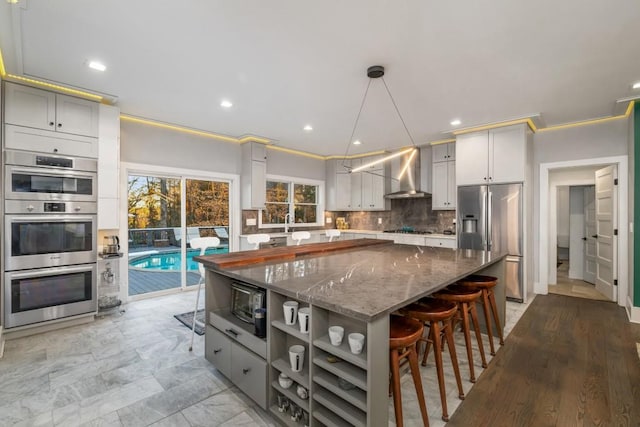 kitchen with hanging light fixtures, stainless steel appliances, a kitchen island, backsplash, and wall chimney exhaust hood