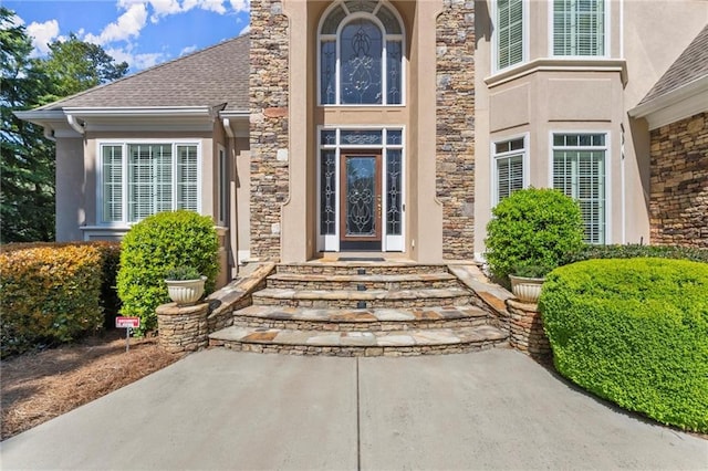 view of exterior entry featuring roof with shingles, stucco siding, and stone siding