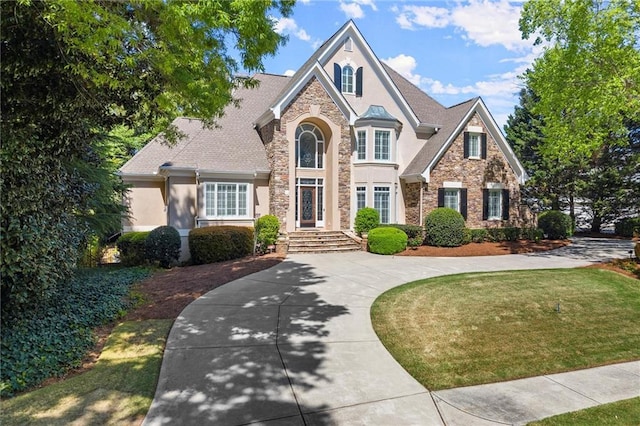 traditional-style home featuring a shingled roof, stone siding, stucco siding, curved driveway, and a front lawn