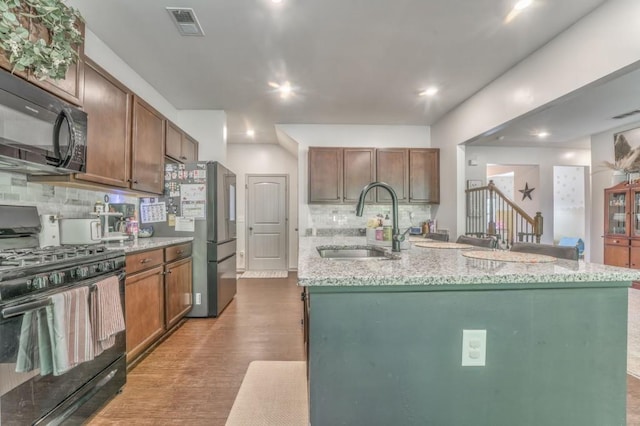 kitchen with black appliances, a sink, backsplash, an island with sink, and light stone countertops