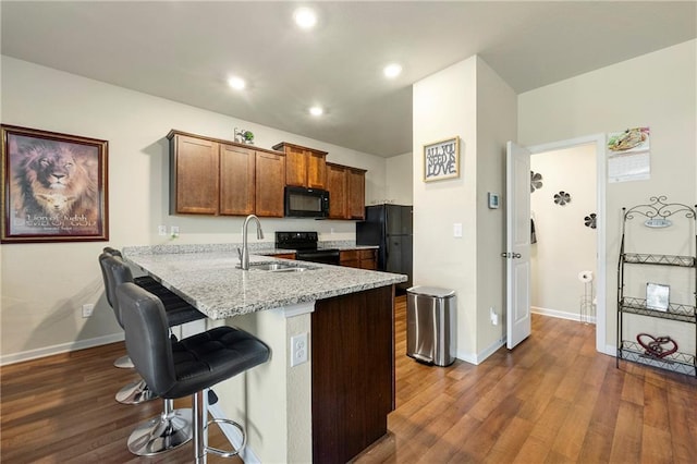 kitchen with light stone counters, dark hardwood / wood-style flooring, sink, black appliances, and a breakfast bar