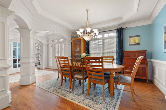 dining space featuring a raised ceiling, crown molding, light wood-type flooring, a notable chandelier, and decorative columns