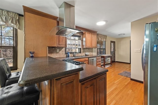 kitchen featuring kitchen peninsula, island exhaust hood, appliances with stainless steel finishes, decorative backsplash, and light hardwood / wood-style flooring