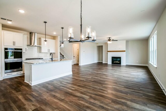 39 Pecan Ct, Hoschton, GA 30548 - kitchen with wall chimney range hood, stainless steel double oven, a chandelier, a ceiling fan, and a glass covered fireplace