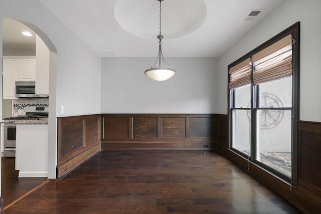 unfurnished dining area with a wainscoted wall, visible vents, arched walkways, dark wood-type flooring, and a raised ceiling