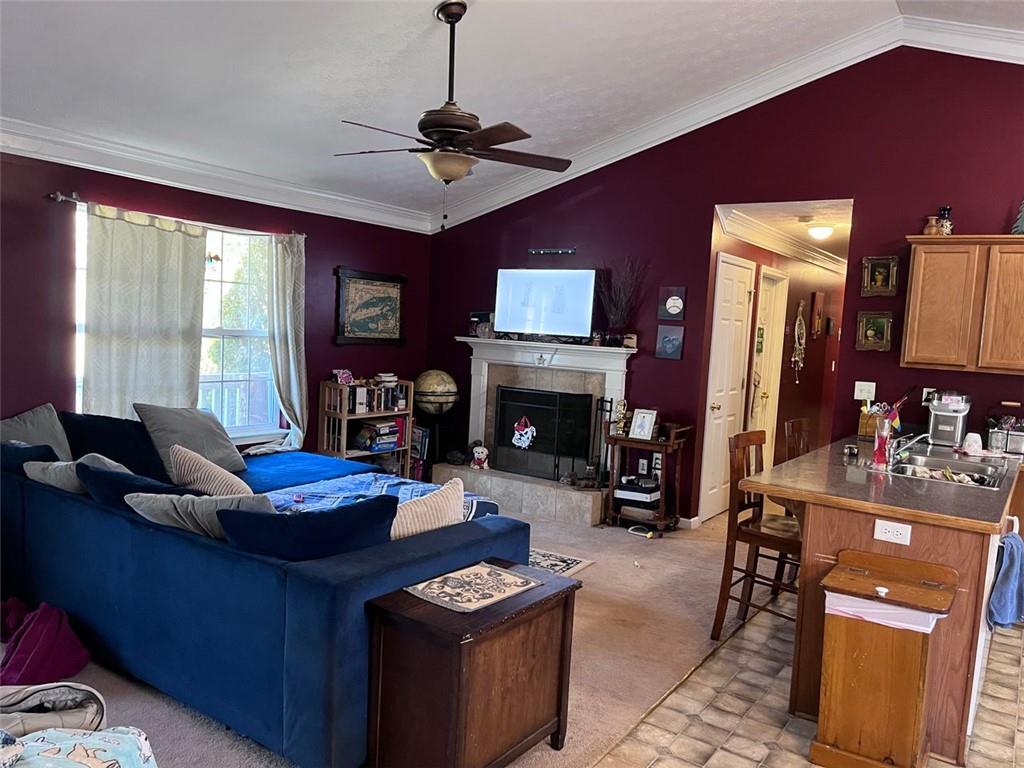 living room featuring a ceiling fan, lofted ceiling, a tile fireplace, ornamental molding, and light colored carpet