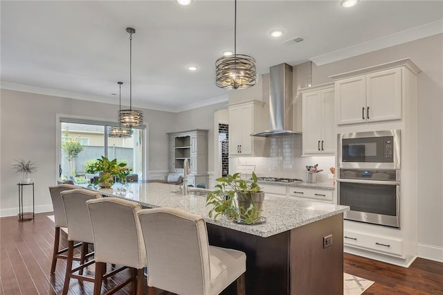 kitchen with appliances with stainless steel finishes, wall chimney exhaust hood, a sink, a kitchen island with sink, and dark wood-style floors