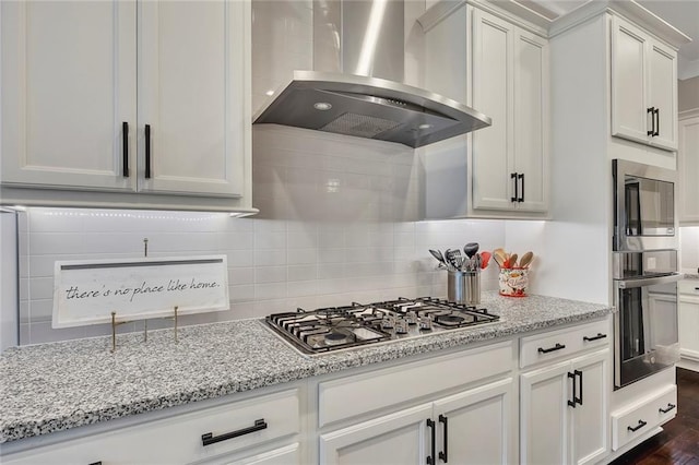 kitchen featuring wall chimney range hood, appliances with stainless steel finishes, white cabinets, light stone countertops, and decorative backsplash