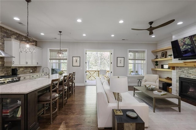 living room with sink, ceiling fan with notable chandelier, hardwood / wood-style floors, a stone fireplace, and crown molding