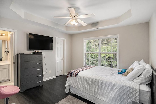 bedroom featuring a raised ceiling, dark wood finished floors, a ceiling fan, ensuite bath, and baseboards