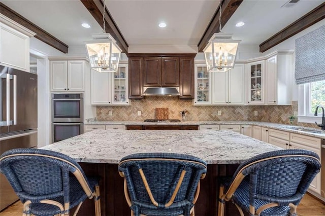 kitchen featuring a chandelier, appliances with stainless steel finishes, a sink, under cabinet range hood, and beamed ceiling