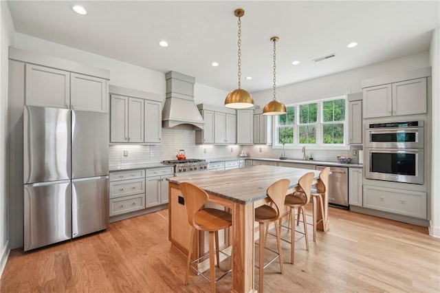 kitchen featuring gray cabinets, backsplash, appliances with stainless steel finishes, light wood-type flooring, and premium range hood