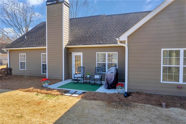 rear view of house featuring a patio area, a lawn, a chimney, and roof with shingles