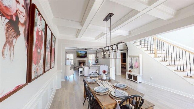 dining room featuring a warm lit fireplace, light wood finished floors, coffered ceiling, stairway, and beam ceiling