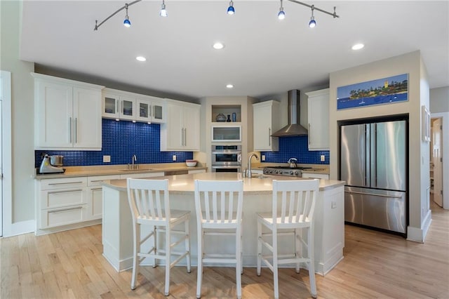 kitchen featuring wall chimney range hood, high end fridge, a sink, decorative backsplash, and a center island with sink