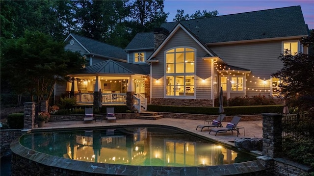 back of house at dusk with a chimney, a patio area, an infinity pool, and stone siding
