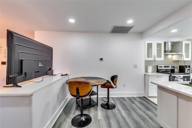 kitchen featuring wall chimney range hood, wood-type flooring, a kitchen breakfast bar, white cabinetry, and stainless steel electric range oven