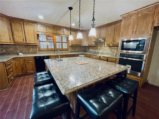 kitchen with brown cabinetry, dark wood-type flooring, a kitchen bar, under cabinet range hood, and black appliances