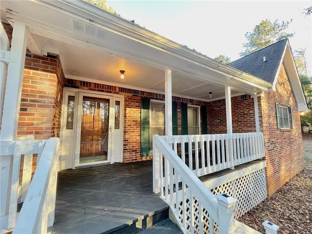 view of exterior entry with roof with shingles, covered porch, and brick siding