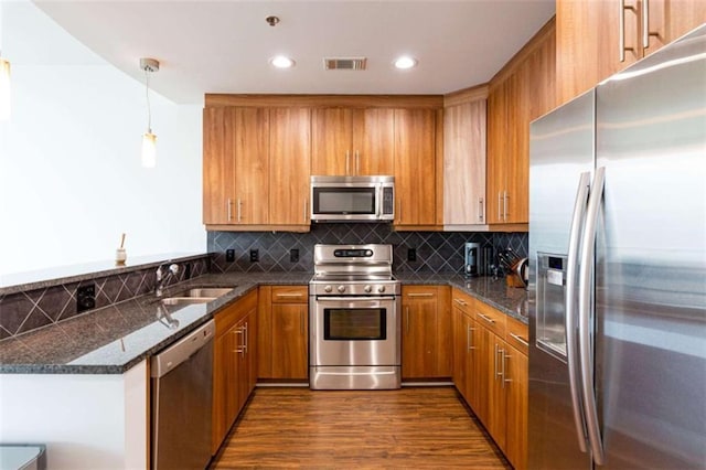 kitchen with stainless steel appliances, a sink, hanging light fixtures, brown cabinets, and dark stone counters