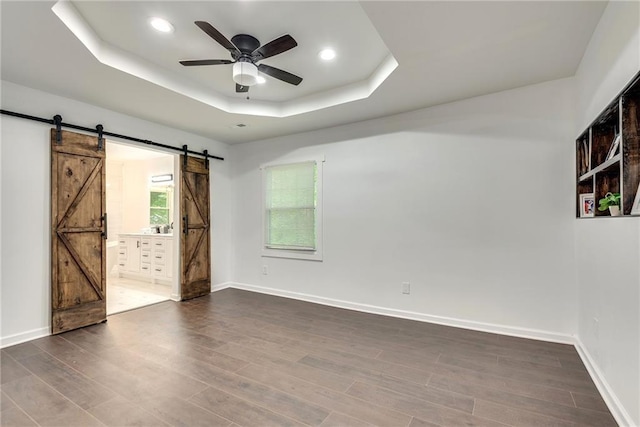 empty room featuring ceiling fan, a tray ceiling, a barn door, and dark hardwood / wood-style flooring