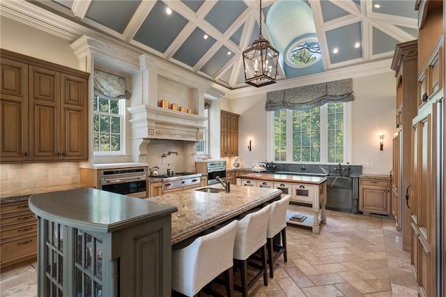 kitchen with coffered ceiling, tasteful backsplash, stainless steel oven, and a kitchen island with sink