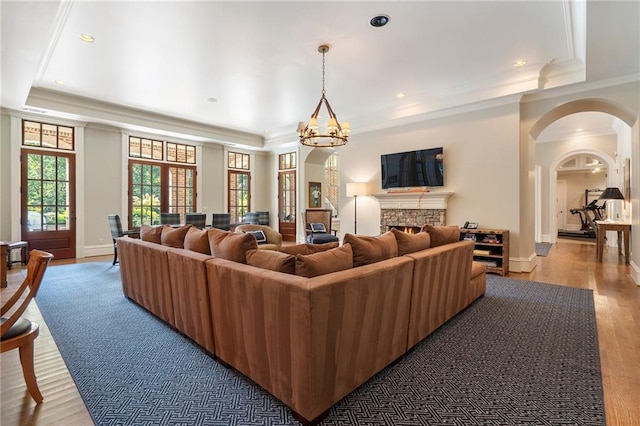 living room with ornamental molding, a tray ceiling, wood finished floors, arched walkways, and a stone fireplace