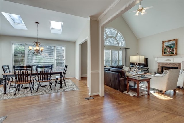 dining room featuring ceiling fan with notable chandelier, light wood-type flooring, high vaulted ceiling, and a brick fireplace