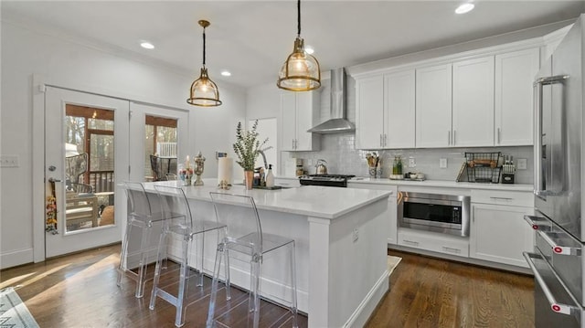 kitchen featuring appliances with stainless steel finishes, wall chimney exhaust hood, a center island with sink, white cabinets, and hanging light fixtures