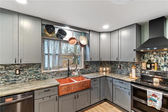 kitchen featuring gray cabinetry, backsplash, stainless steel appliances, light stone counters, and wall chimney exhaust hood