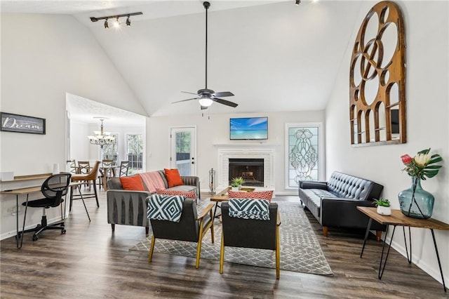 living room featuring a brick fireplace, rail lighting, wood finished floors, baseboards, and ceiling fan with notable chandelier