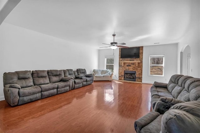 living area with arched walkways, ceiling fan, wood finished floors, a stone fireplace, and baseboards