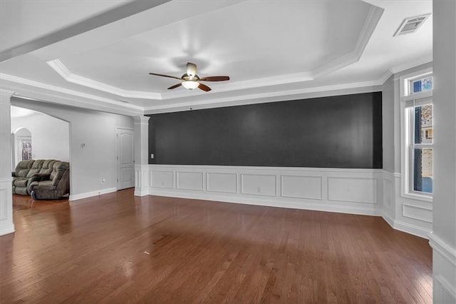 empty room with a tray ceiling, a ceiling fan, dark wood finished floors, ornamental molding, and a decorative wall
