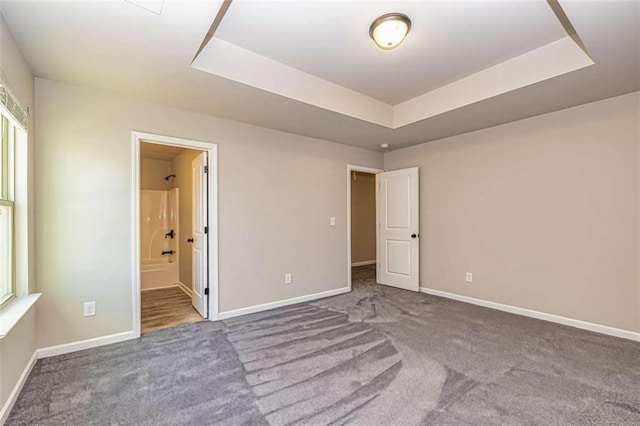 unfurnished bedroom featuring a raised ceiling, baseboards, ensuite bath, and carpet floors