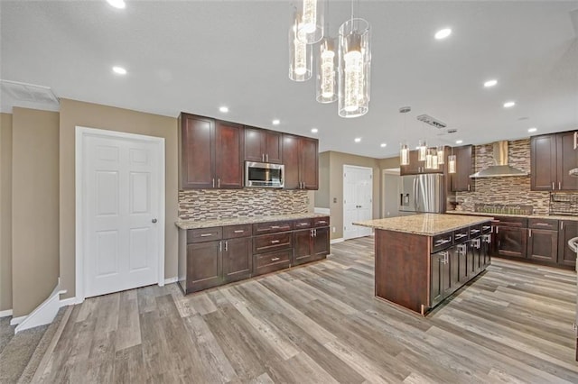 kitchen with stainless steel appliances, hanging light fixtures, a kitchen island, wall chimney range hood, and light wood-type flooring