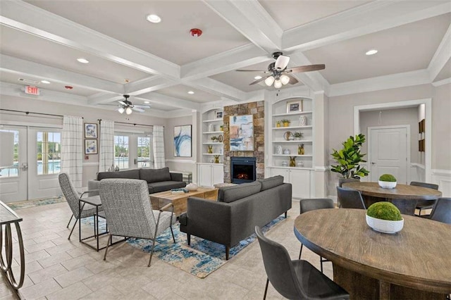 living area with built in shelves, beam ceiling, a stone fireplace, coffered ceiling, and a ceiling fan
