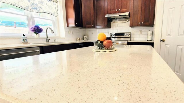 kitchen featuring light stone counters, under cabinet range hood, stainless steel appliances, a sink, and tasteful backsplash